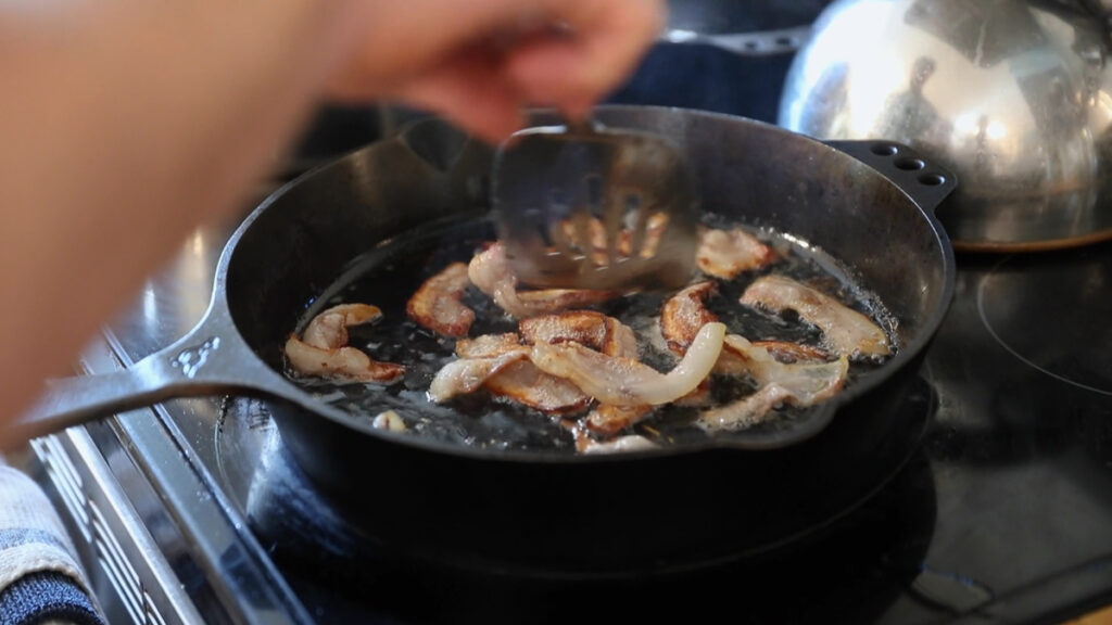 Bacon frying up in a cast iron pan.