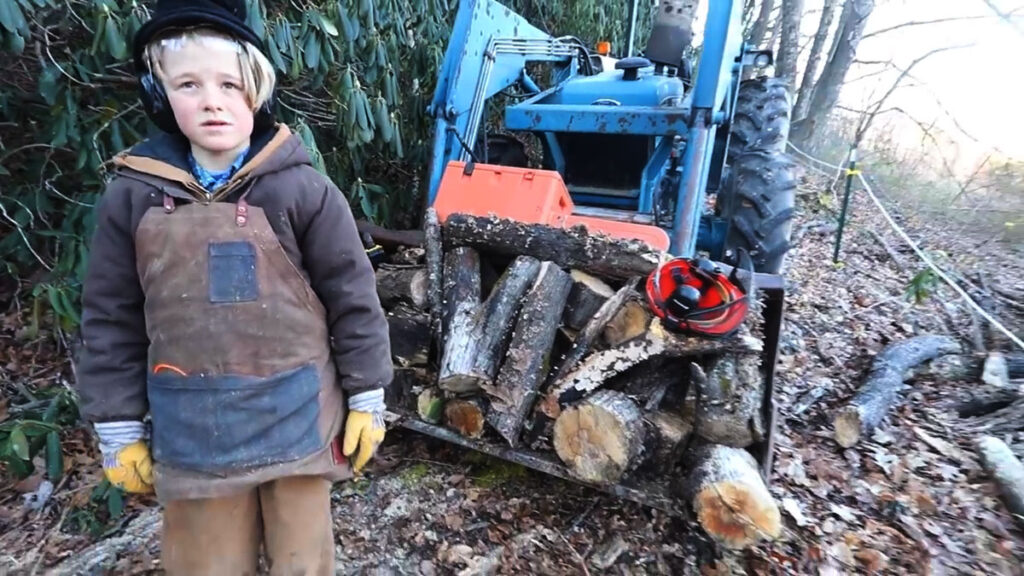 A boy standing by a tractor loaded with rotting logs.