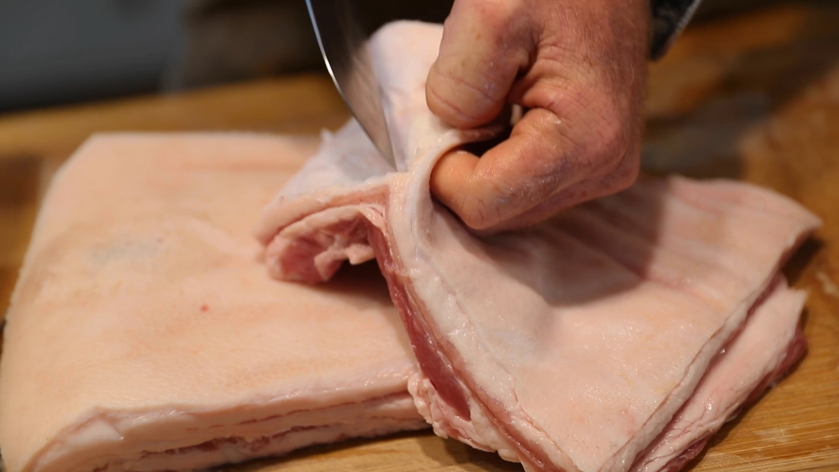 A knife cutting of the pork skin from a slab of pork belly.