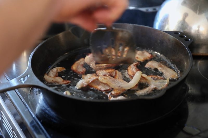 Bacon frying up in a cast iron pan.